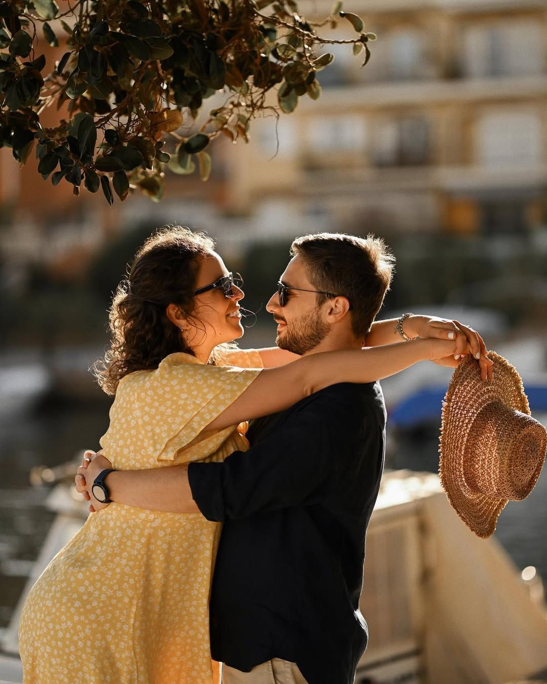 A woman in a vibrant yellow dress holds a sun hat while hugging a man against the backdrop of colorful buildings in Port Saplaya