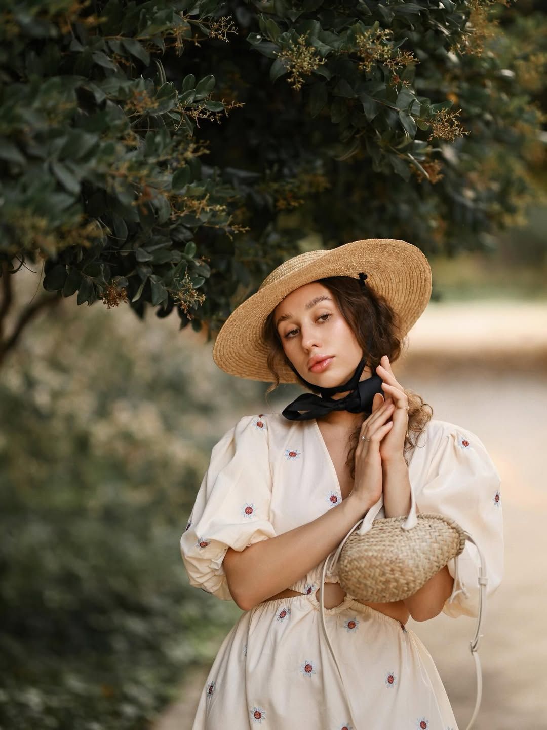 A beautiful women's portrait of a lady in a straw hat with a black ribbon, posing elegantly under lush green trees in a Valencia park.