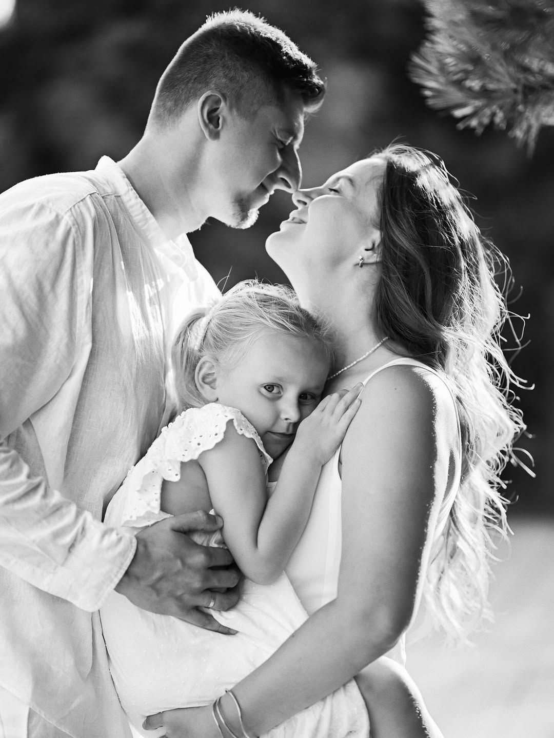 An intimate black and white portrait showing a father kissing his wife's forehead while holding their daughter.