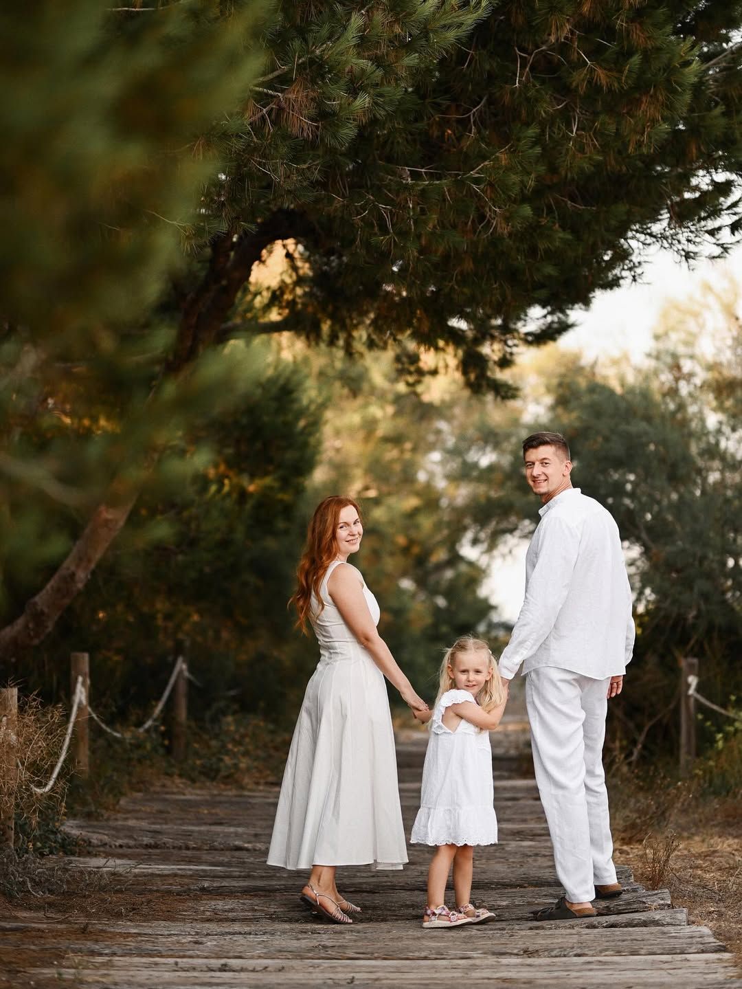 A family walking on a wooden boardwalk among pine trees in El Saler, with mom and daughter looking at the camera.