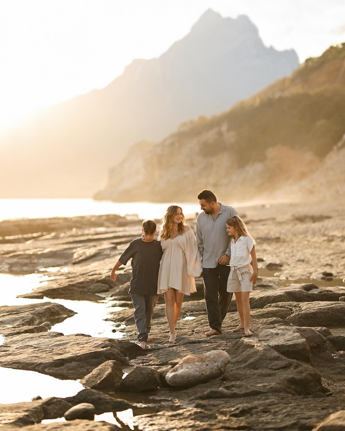 A family with two children walking hand in hand along a rocky shoreline with a mountain in the background in Costa Blanca.