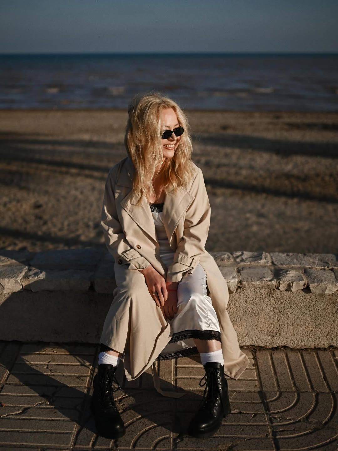 A smiling blonde woman in a trench coat and black boots sitting on a stone promenade by the beach in Costa Blanca.