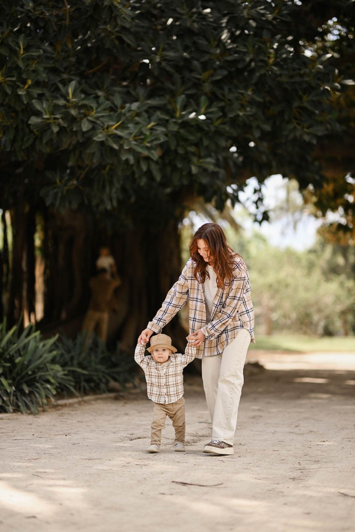 A mother holds her toddler son's hands to help him walk along a wide dirt path under the large trees of Viveros Park.