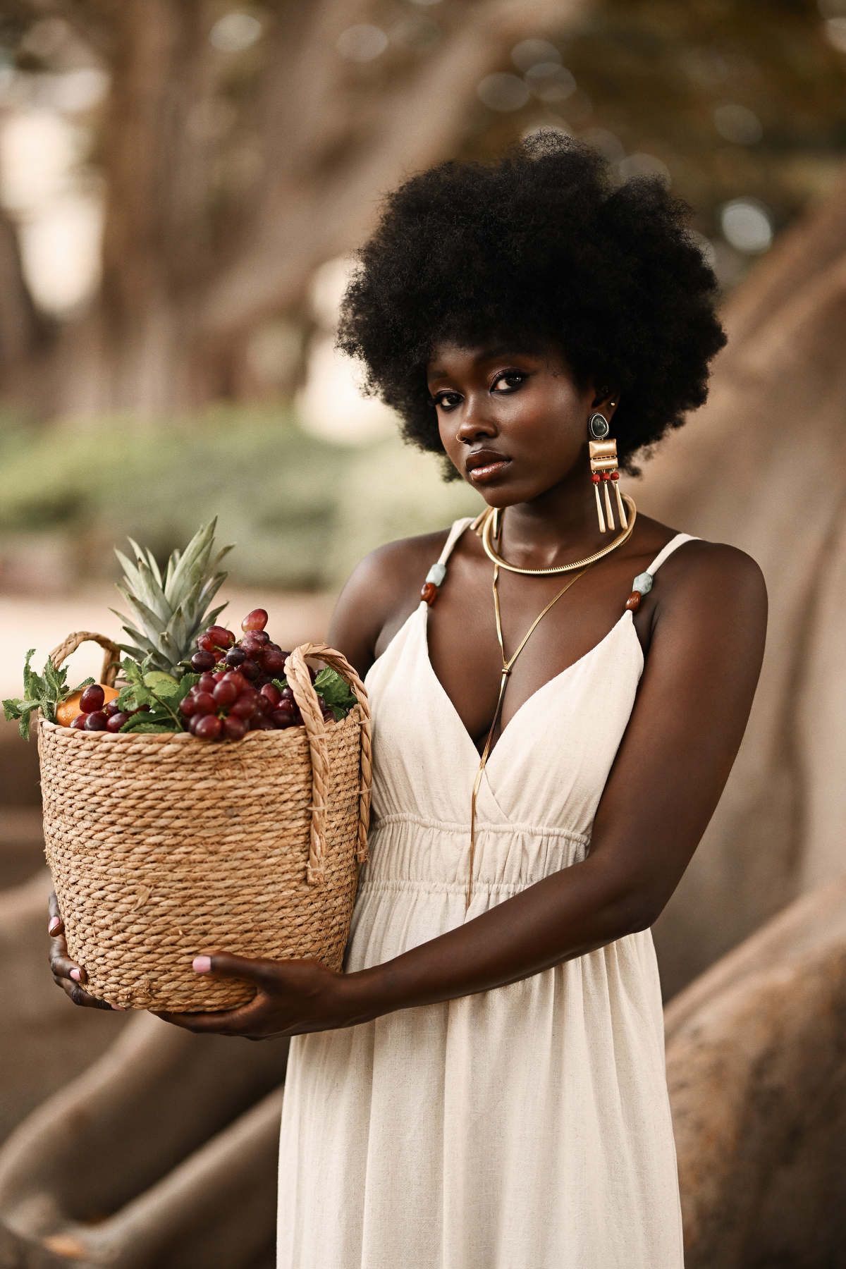 Editorial portrait of a woman in a cream dress holding a fruit basket against a textured tree backdrop at La Glorieta park.