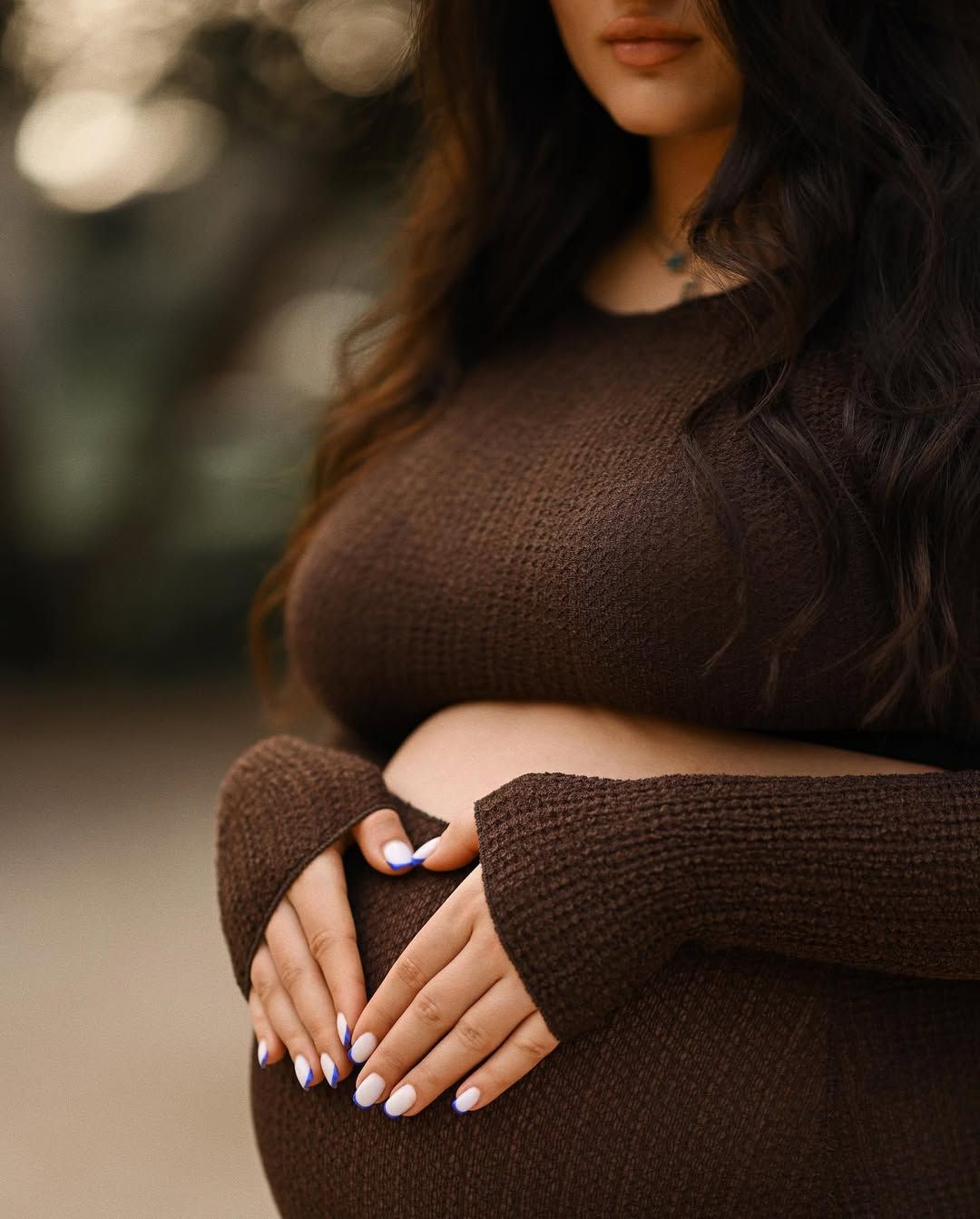 Close-up of a mother's hands resting on her pregnant belly wearing a brown fitted dress during a maternity session in Valencia.