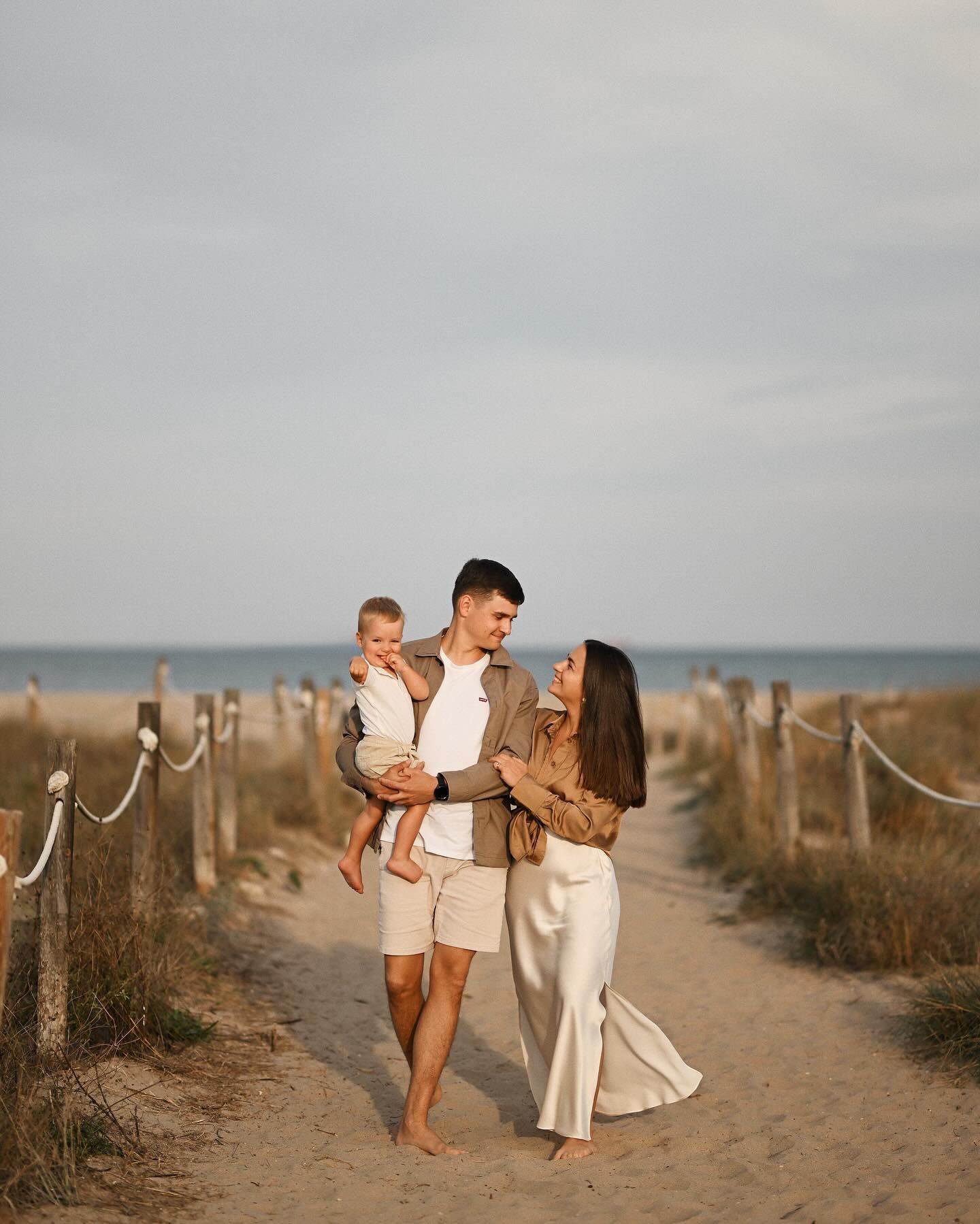 Smiling parents walking along a sandy beach pathway while carrying their small child during a lifestyle family photoshoot in Valencia.