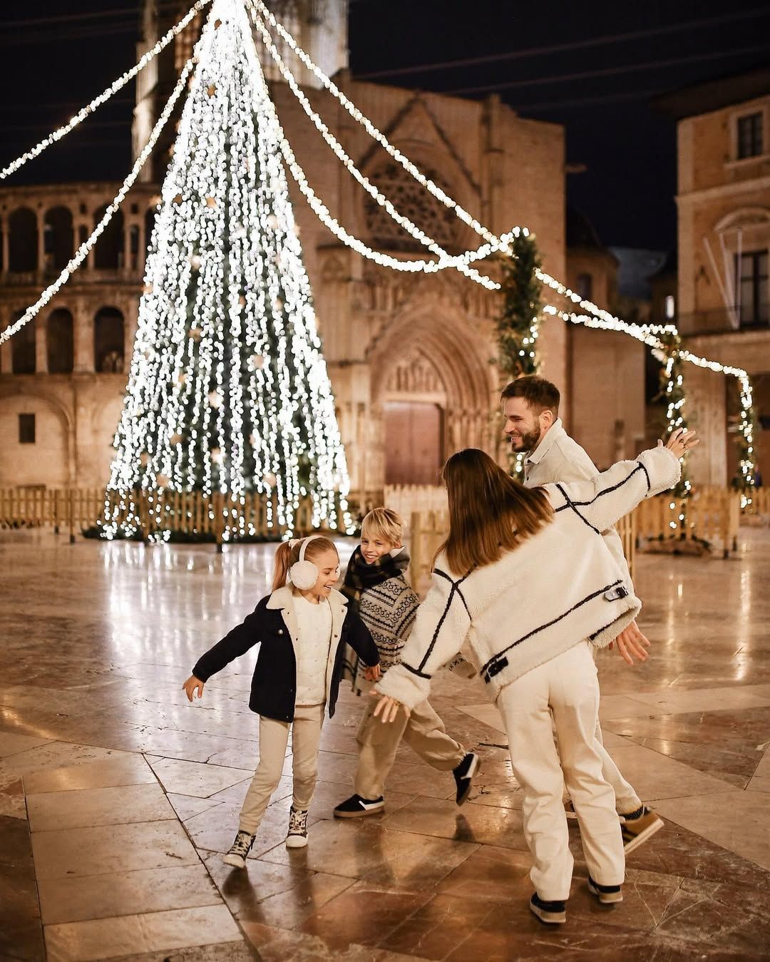 Playful family holding hands and dancing near a giant lit Christmas tree in a historic Valencia square at night