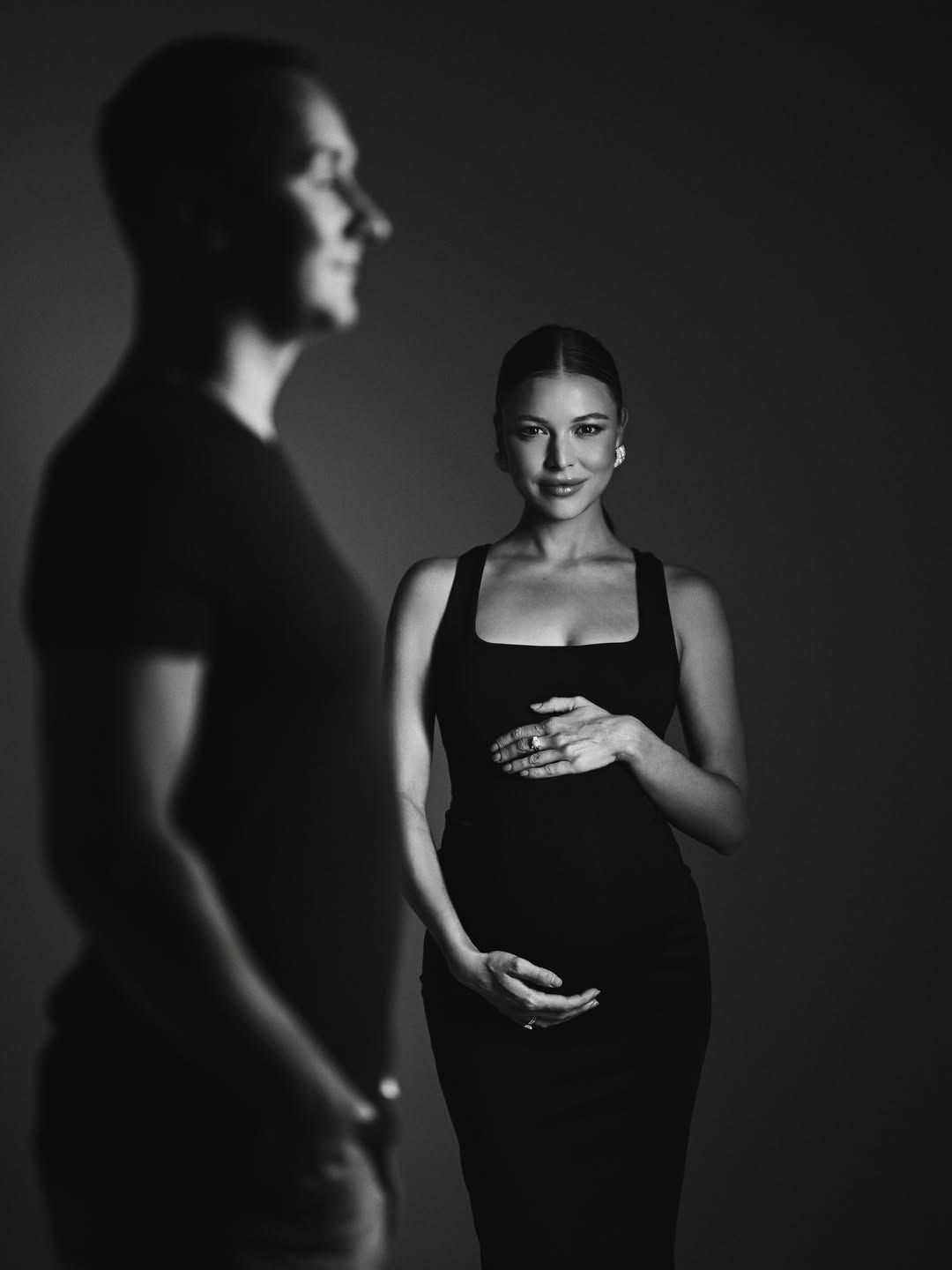 Elegant black and white studio portrait of a pregnant woman in a black dress with her partner in the foreground
