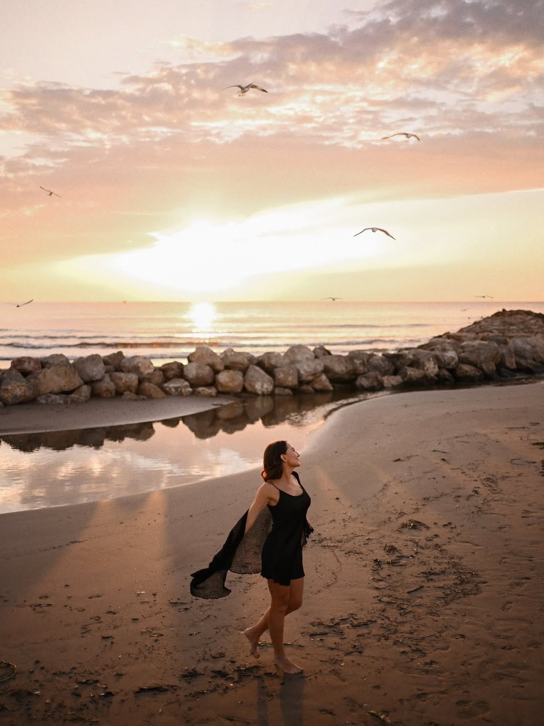 Female lifestyle session on a Valencia beach, walking on the sand with a dark shawl under a sunset sky with flying seagulls.