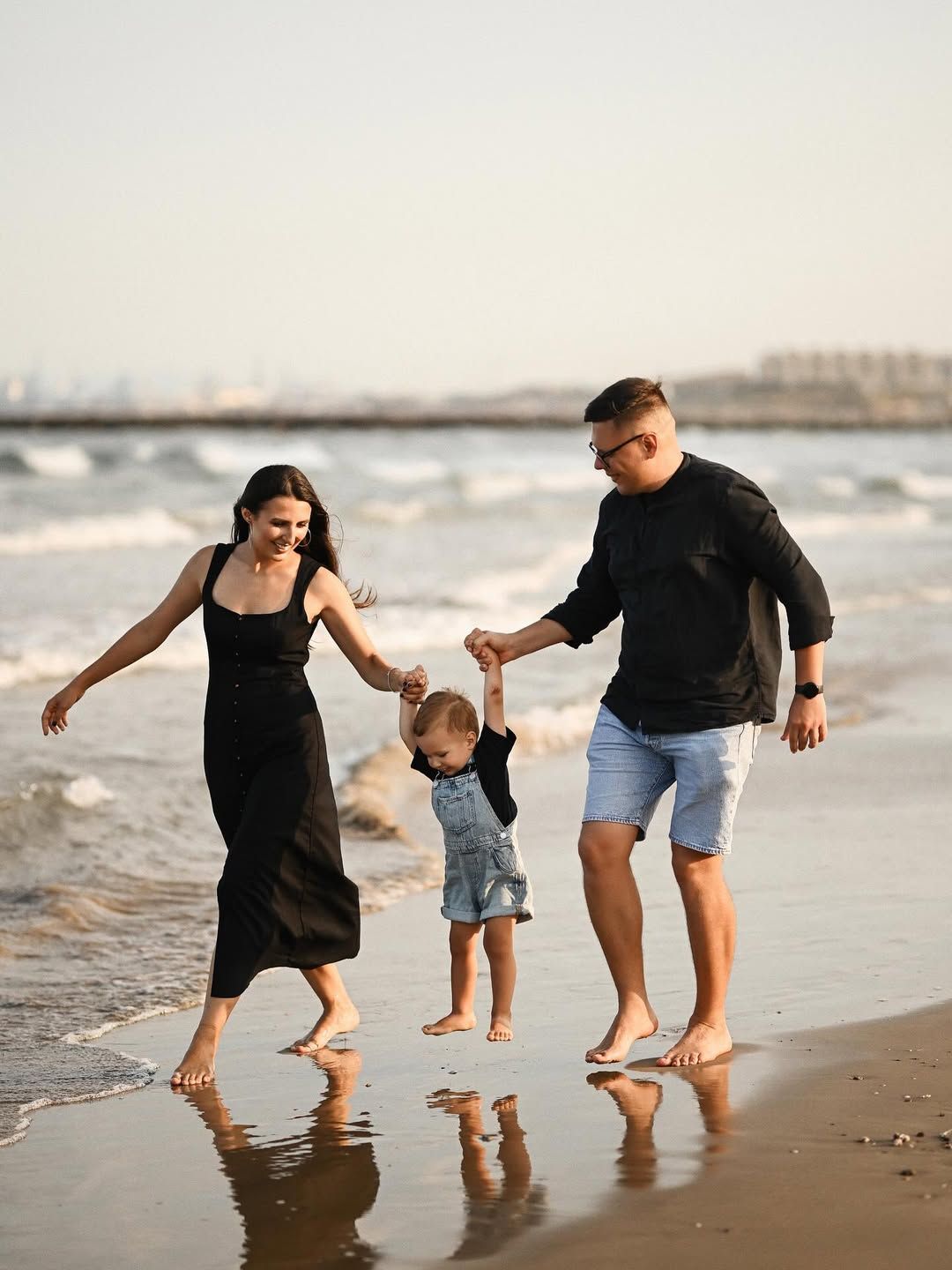 A family of three walks along the sandy shore with rolling waves on the Mediterranean beach in Port Saplaya, Valencia