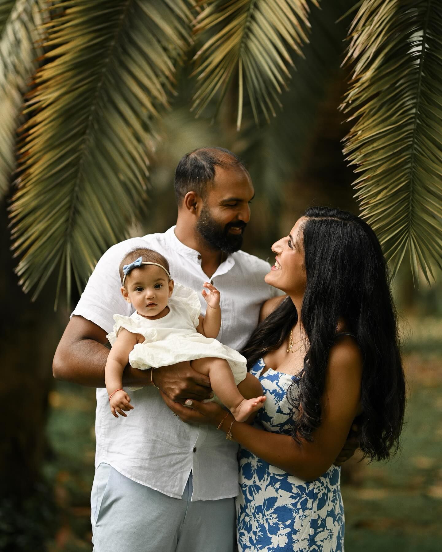 A happy couple sharing a tender moment while holding their baby daughter outdoors in a park, framed by beautiful tropical palm fronds.
