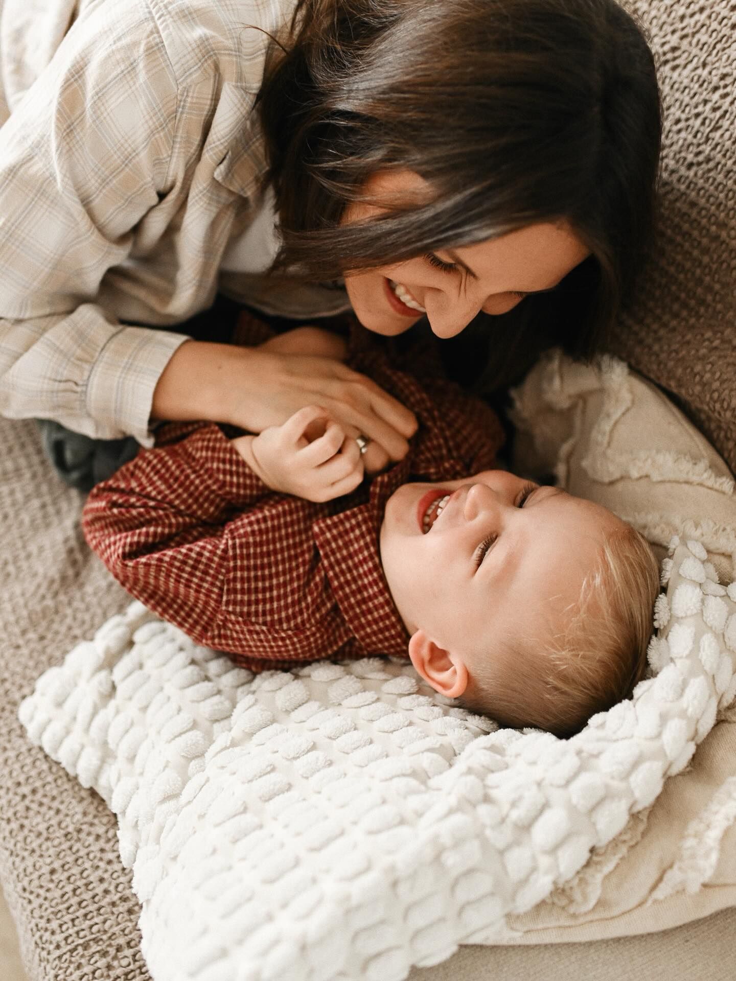A joyful mother laughing and cuddling with her young son on a cozy sofa, capturing an intimate and genuine moment during a home photoshoot.