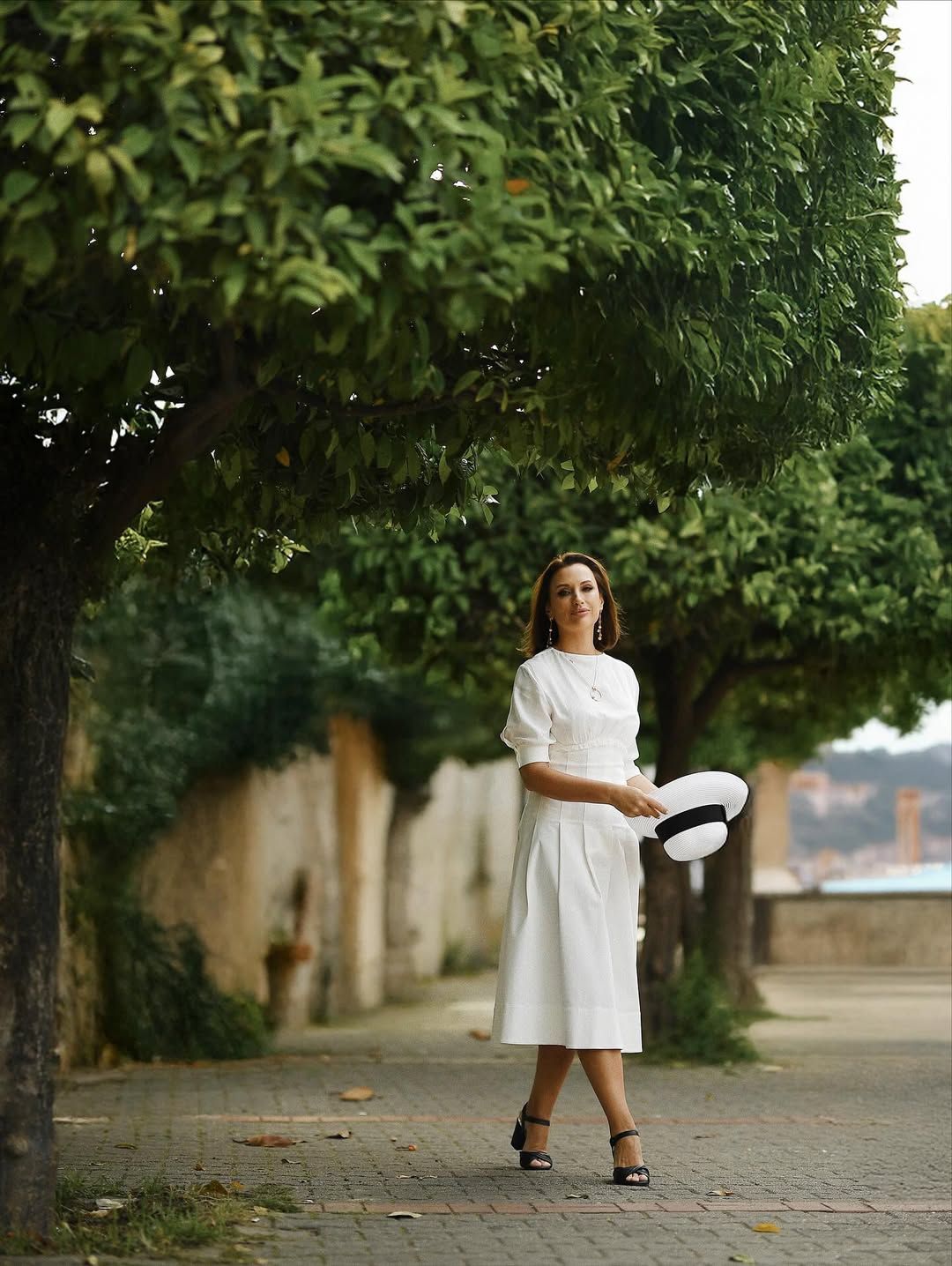 A woman in a white dress walking down a tree-lined pavement, holding a straw hat during a lifestyle portrait session in Valencia.