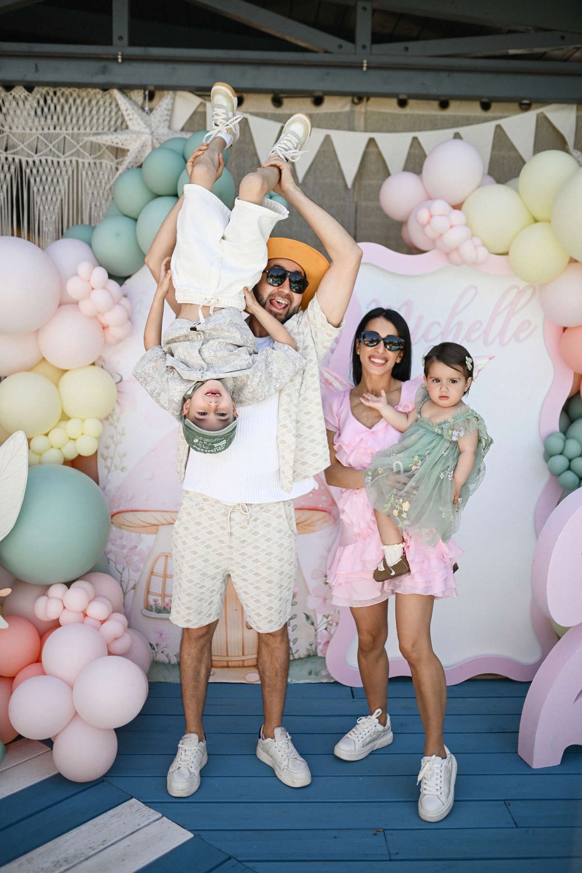 A playful father holding his young son upside down next to his wife and daughter at an outdoor birthday celebration.