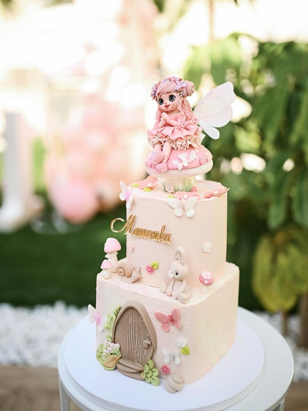 Elegant two-tier pink birthday cake decorated with a miniature fairy figurine, butterflies, and a tiny door.
