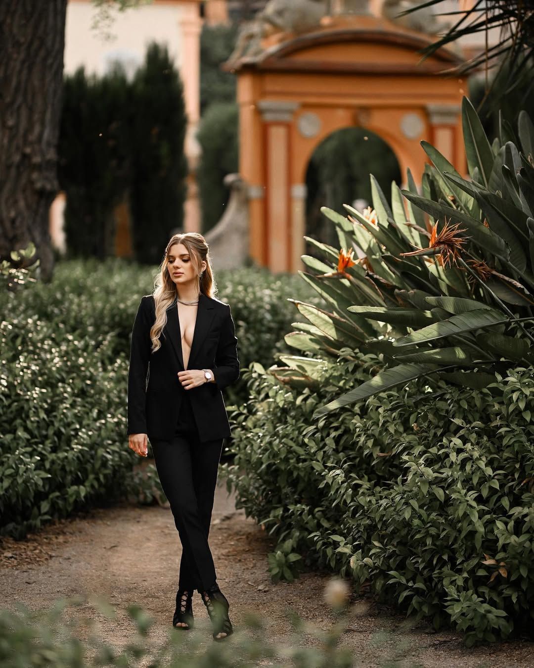 A woman posing in a black suit on a path next to tropical plants and a historic orange arch in Valencia.