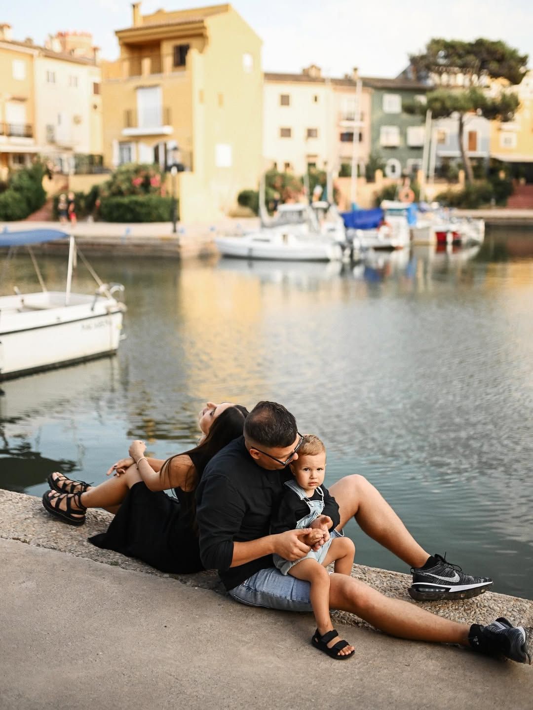 A family sits together on a wooden pier with a beautiful backdrop of traditional boats and colorful houses in Port Saplaya