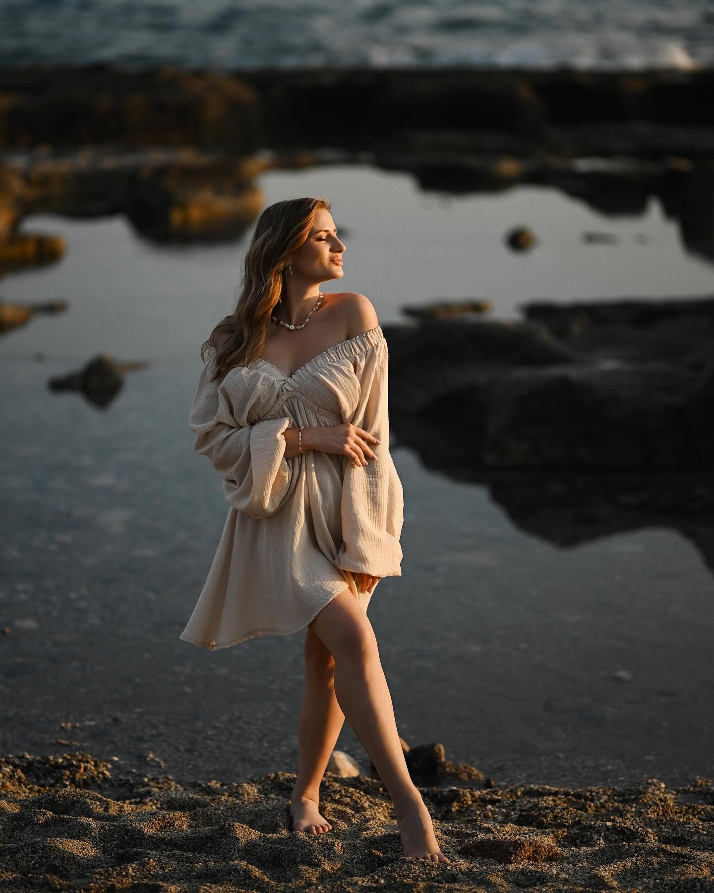 Full-length portrait of a pregnant woman in an off-the-shoulder beige dress standing by tidal pools in Costa Blanca.