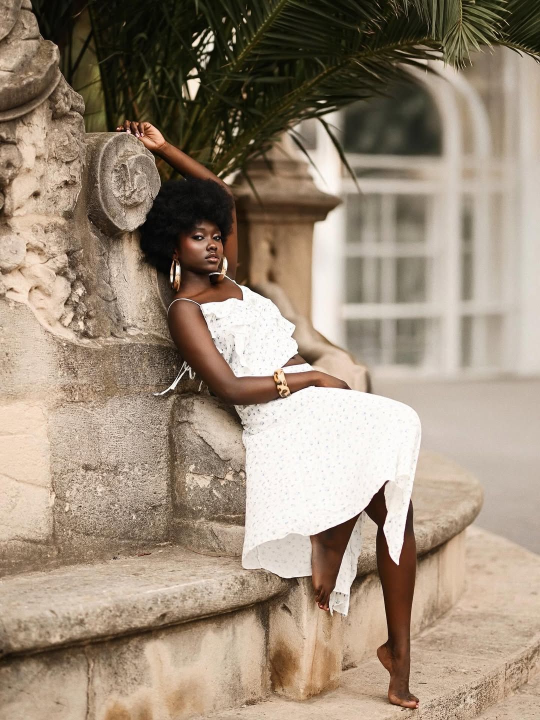 Full-length portrait of a model leaning against an ancient stone fountain under palm trees during a photoshoot in Alicante.