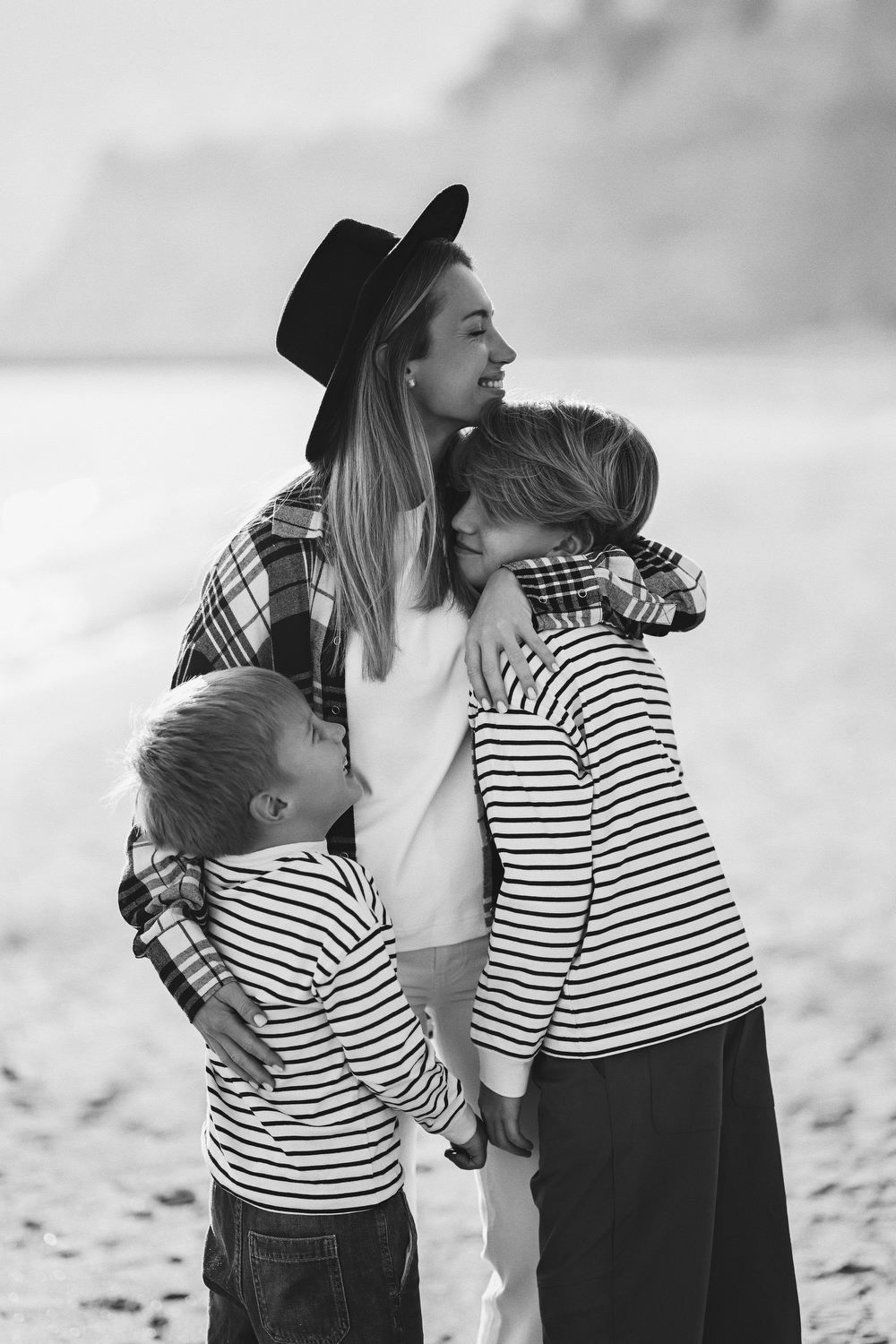 Candid black and white portrait of a smiling mother lovingly embracing her two sons on the beach during a session in Calpe