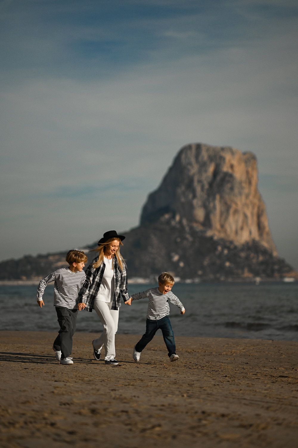 A happy family holding hands and running on the sandy beach with the iconic Penyal d'Ifac mountain in the background in Calpe