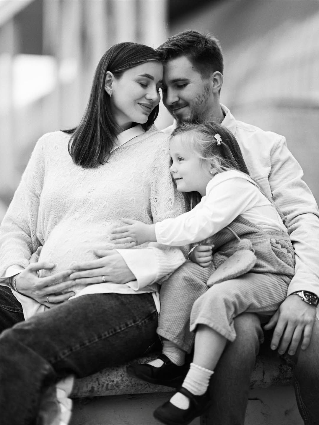 Black and white maternity portrait in Turia Park, Valencia with parents embracing as their daughter gently touches the baby bump.