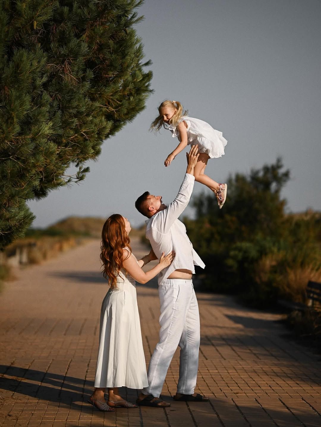 A father tossing his laughing daughter in a white dress into the air along a pine-lined path in El Saler.