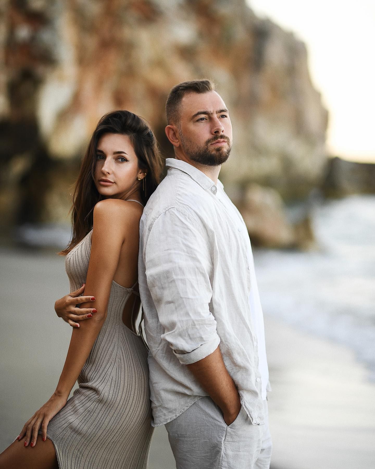 A cinematic portrait of a beautiful couple standing back-to-back on a serene Valencia beach against dramatic rocky coastal cliffs.