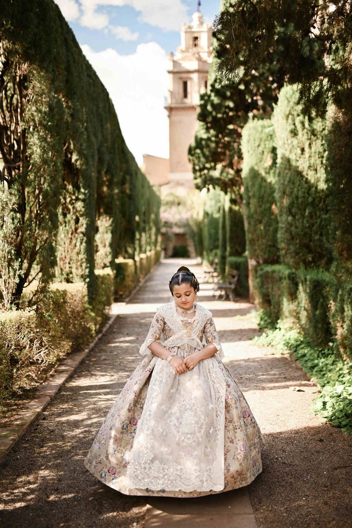 A graceful full-length portrait of a young girl standing elegantly along a cypress-lined path in the neoclassical Jardines de Monforte.