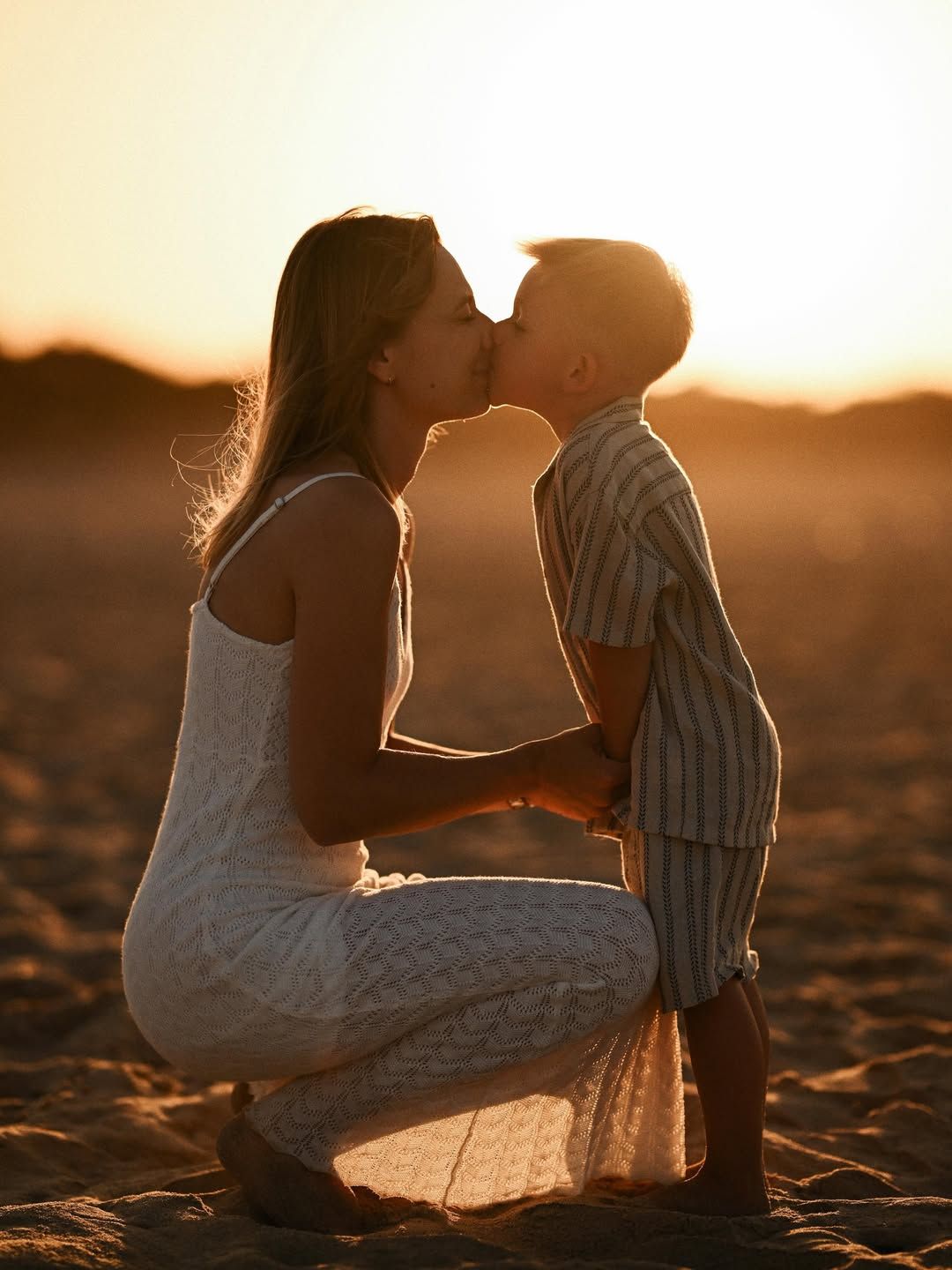 A tender sunset portrait of a mother and her son sharing a sweet kiss on the sandy beach of El Saler in Valencia.