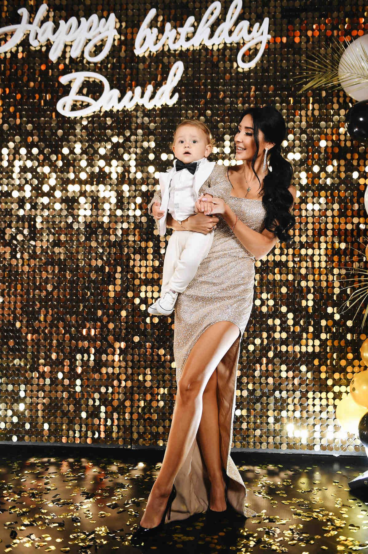 Glamorous gold sequin photo zone with a mother holding her baby boy in a white tuxedo for his first birthday party.