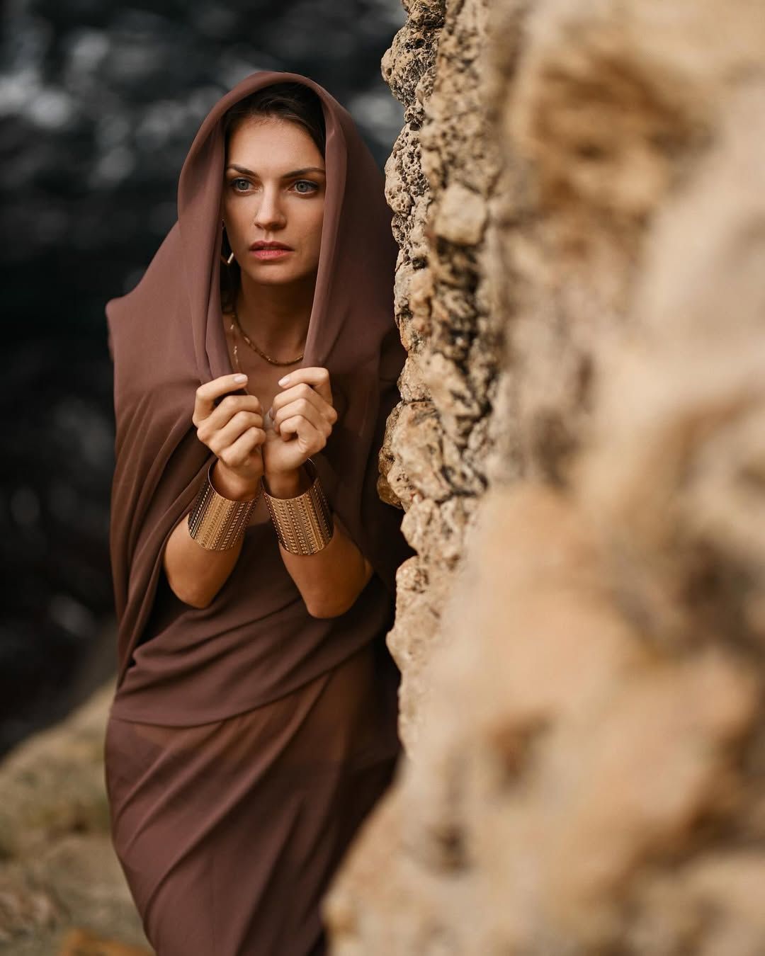 A woman with a draped fabric hood over her head posing against a textured limestone cliff at Cala Moraig.