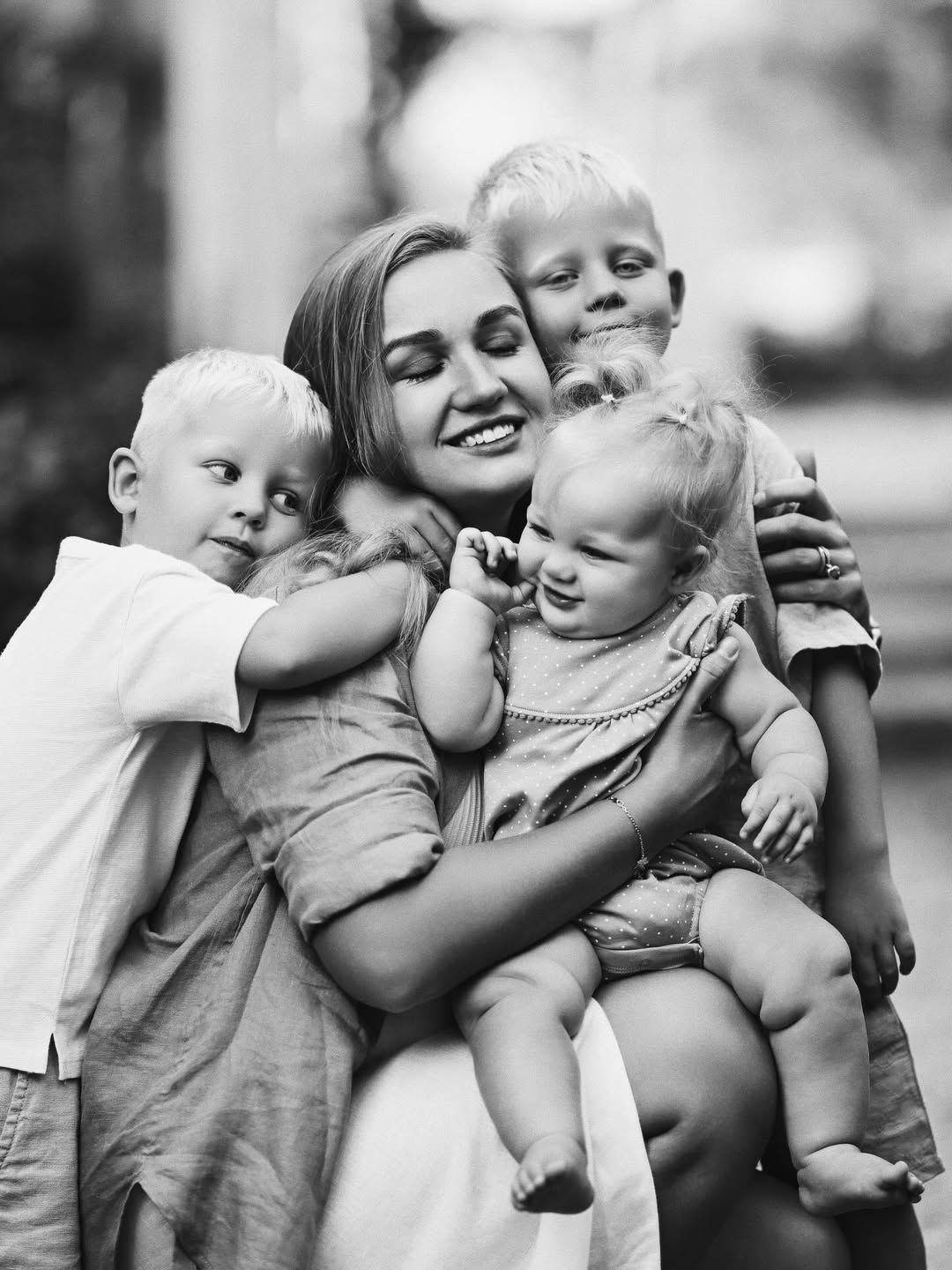 A candid black and white portrait of a mother embracing her three children in Turia Gardens, Valencia.