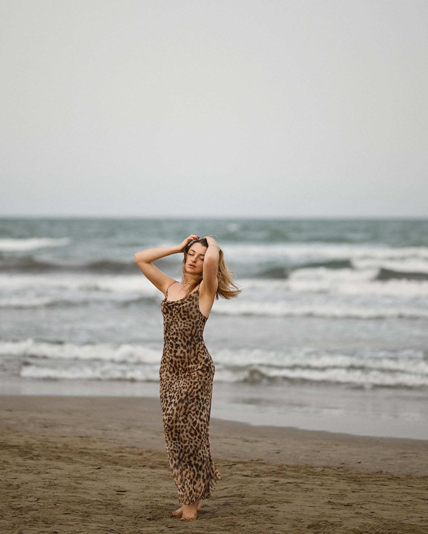 A woman in a long leopard dress posing on a sandy shoreline with blue sea waves in the background during an Alicante photoshoot.