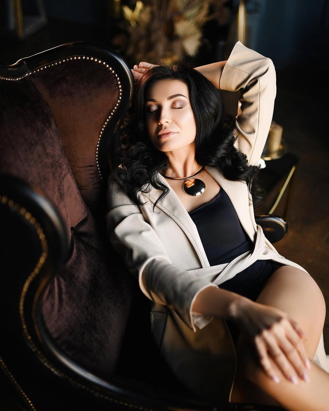 Elegant boudoir photography in Valencia. A brunette woman relaxes in a classic armchair wearing a beige blazer over a black bodysuit.