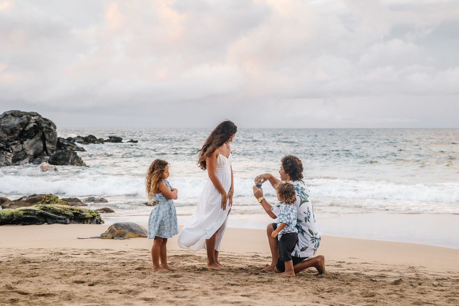 A family enjoys a peaceful moment together on a rocky beach at sunset with waves crashing in the background.