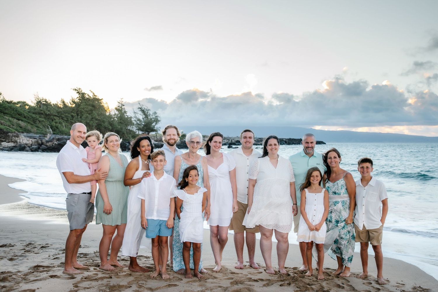 Large family group portrait on a tropical beach at sunset, wearing white and casual outfits, smiling together.