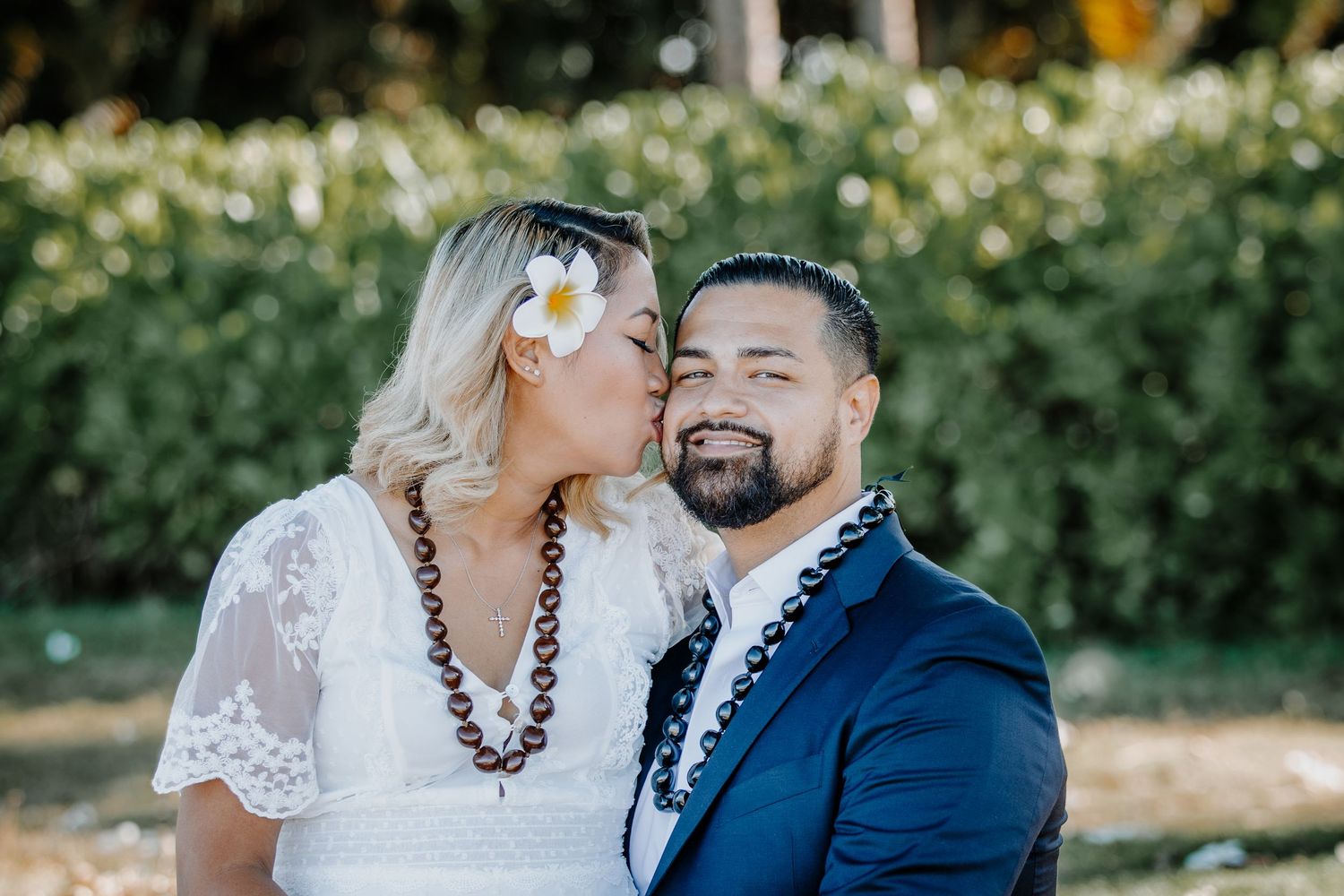 Bride with flower in hair kisses groom in blue suit outdoors at tropical wedding ceremony.
