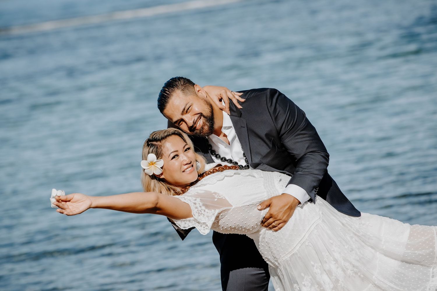 Bride and groom embrace joyfully on a beach, with the groom dipping the bride in a white wedding dress near blue water.