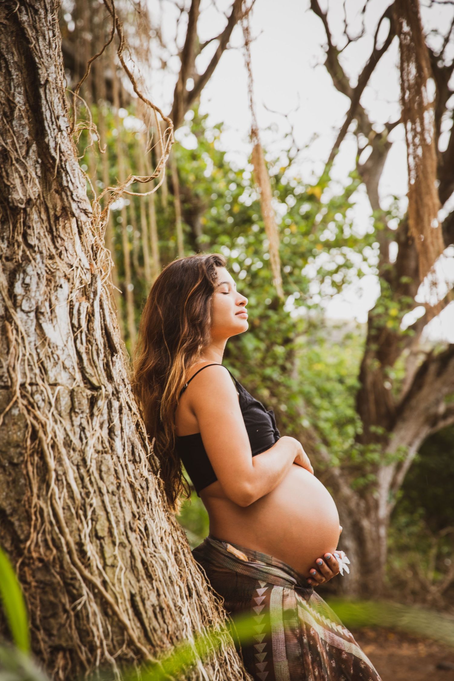 Maternity photo of expectant mother leaning against tree trunk in natural setting.
