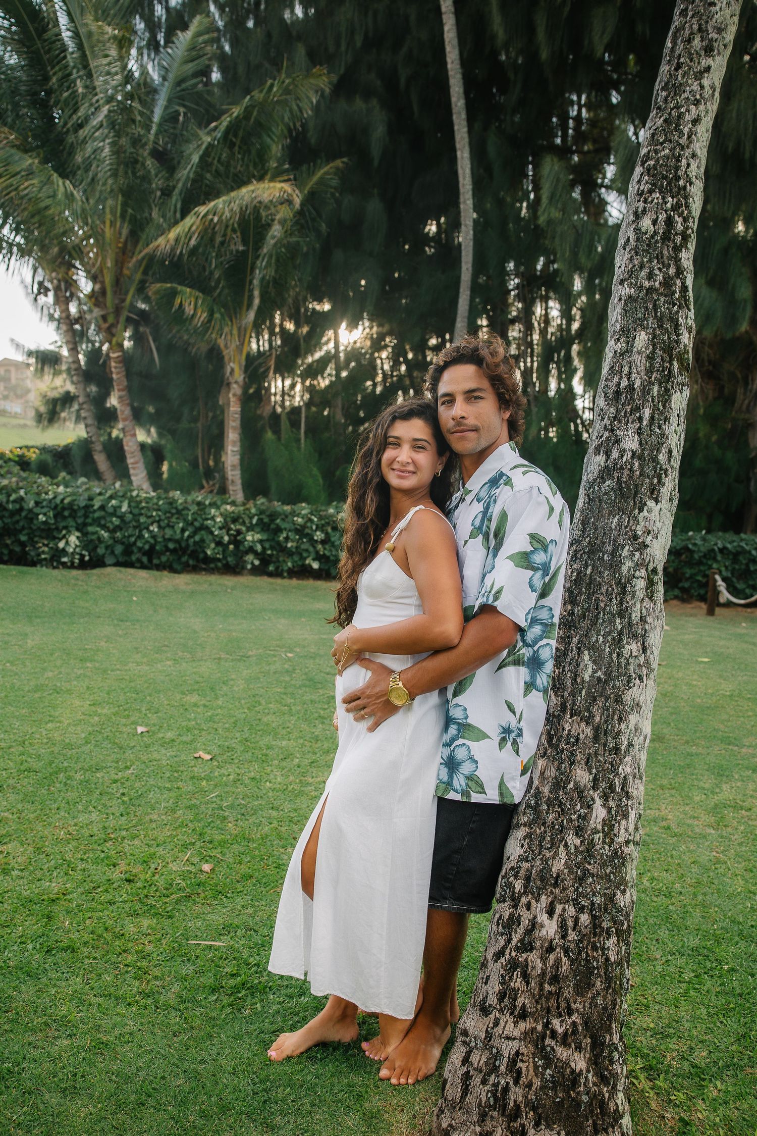 Couple embraces by palm trees wearing white dress and floral shirt in tropical setting.