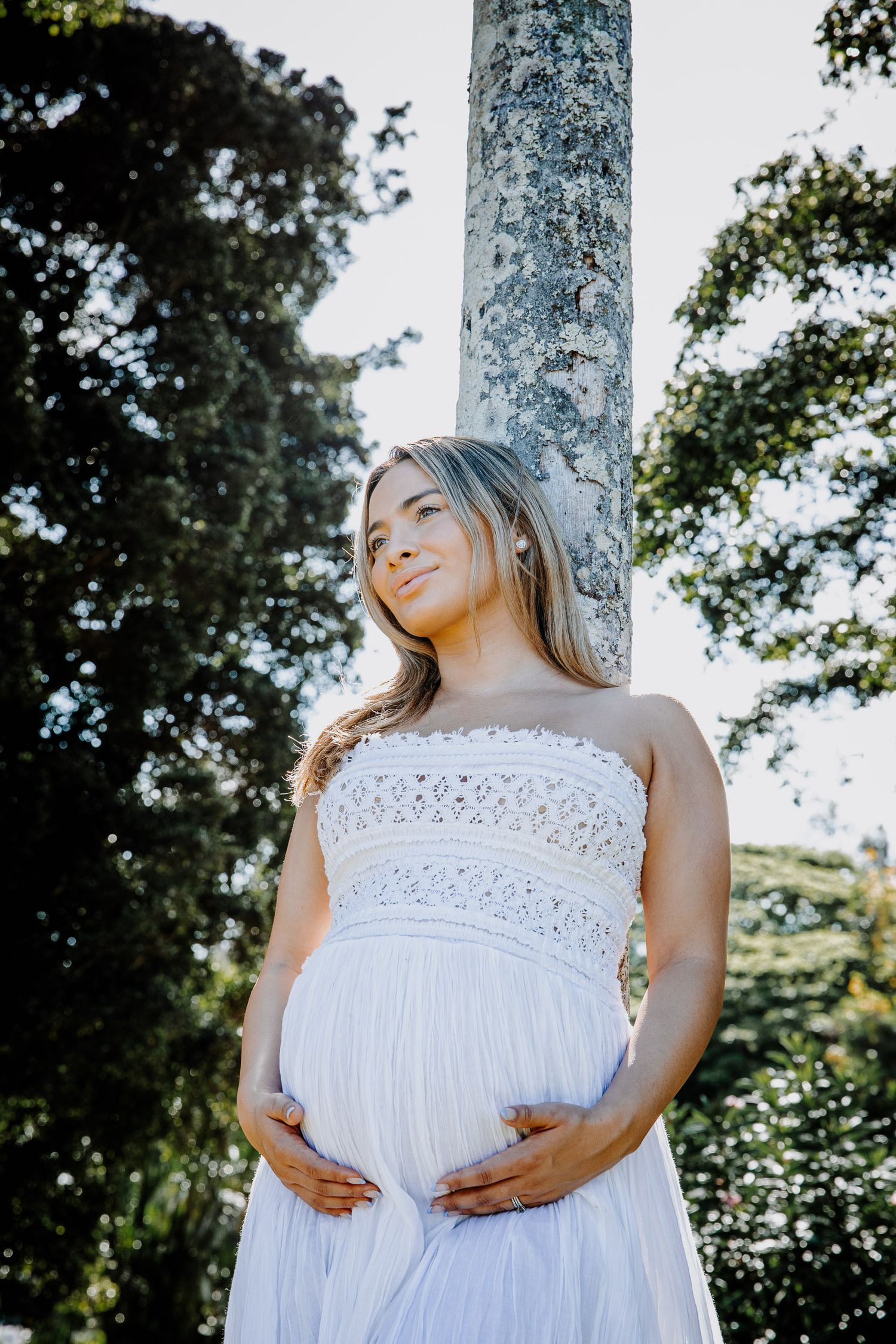 A maternity photo of a woman in a white lace dress standing by a stone pillar in a garden setting.