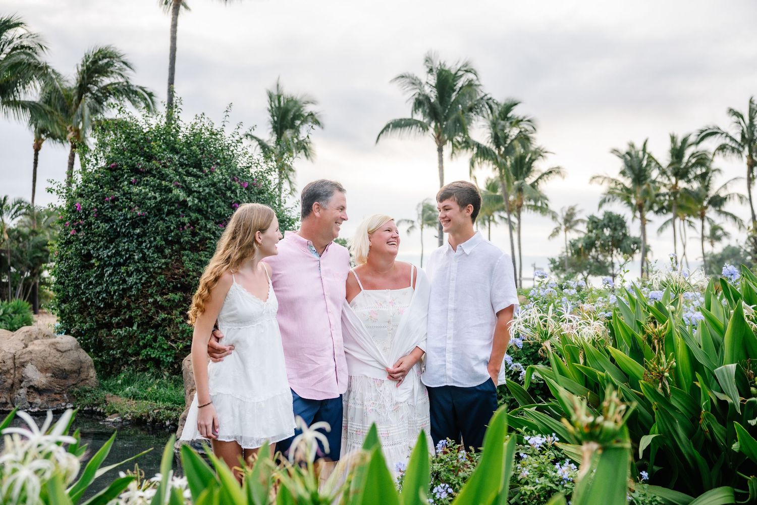 Family of four smiling together in a lush tropical garden with palm trees and white flowers.