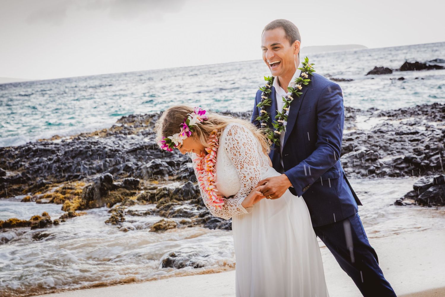 Bride and groom share a joyful moment on a rocky Hawaiian beach, with lei and floral crown.