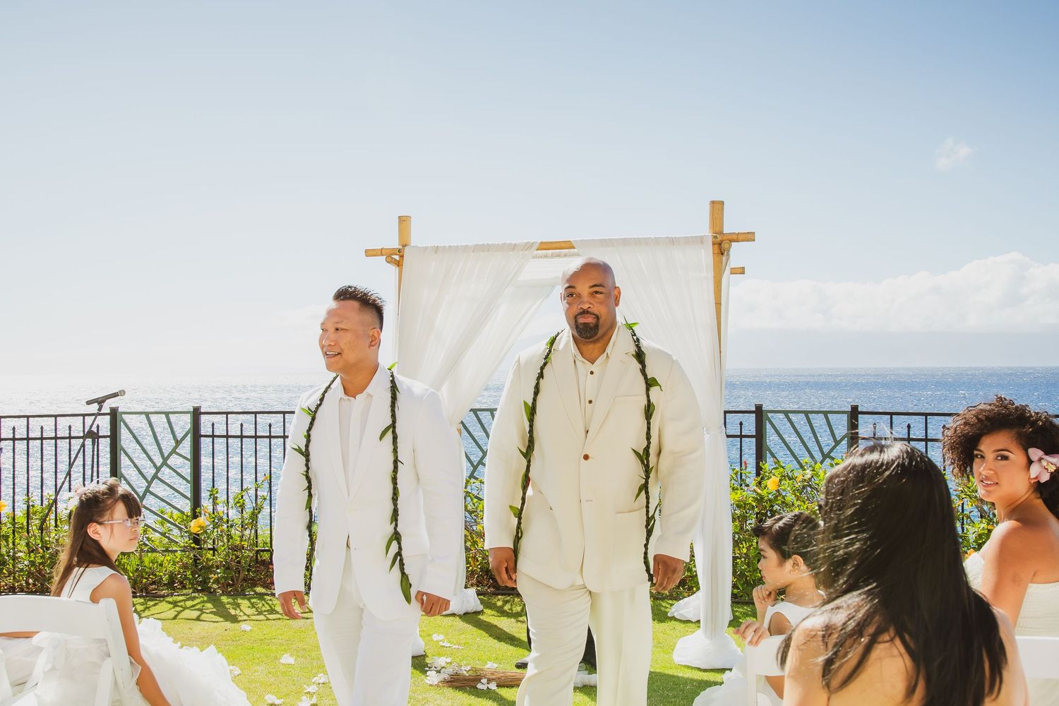 Two grooms in white suits stand at an outdoor wedding altar with ocean views in Hawaii.