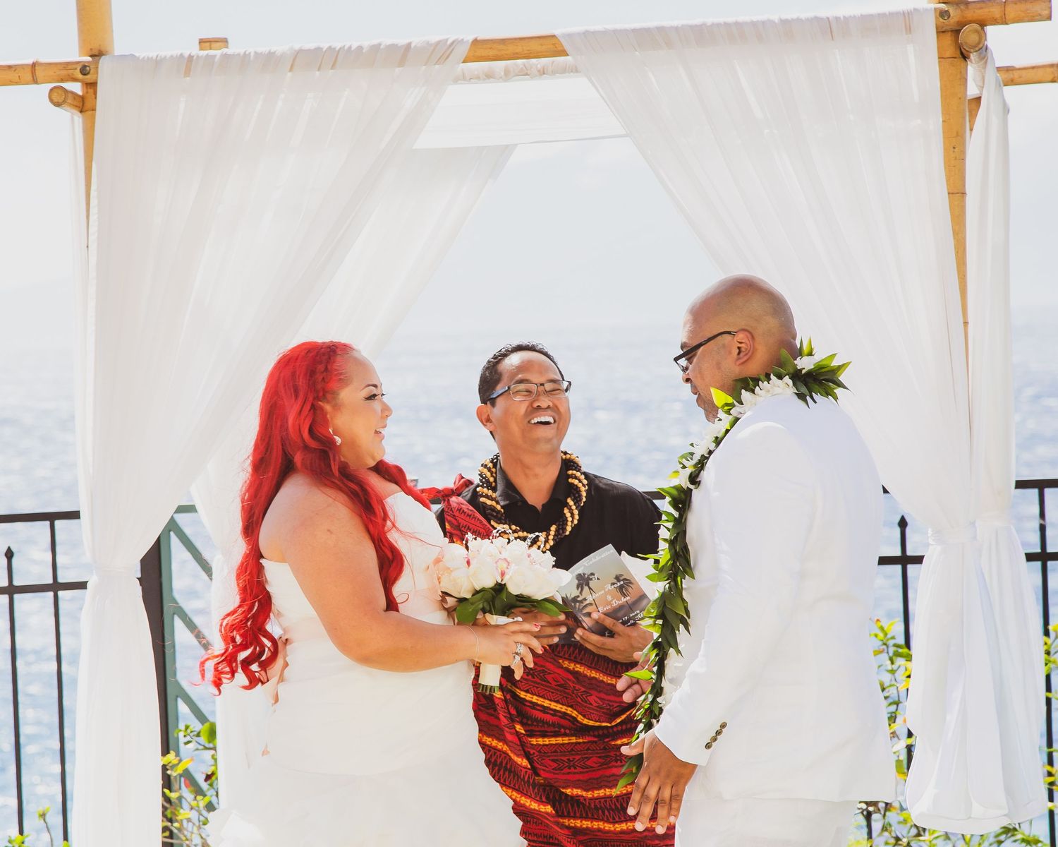 Joyful outdoor wedding ceremony with bride in white dress, groom in white suit, and officiant under a draped arch.