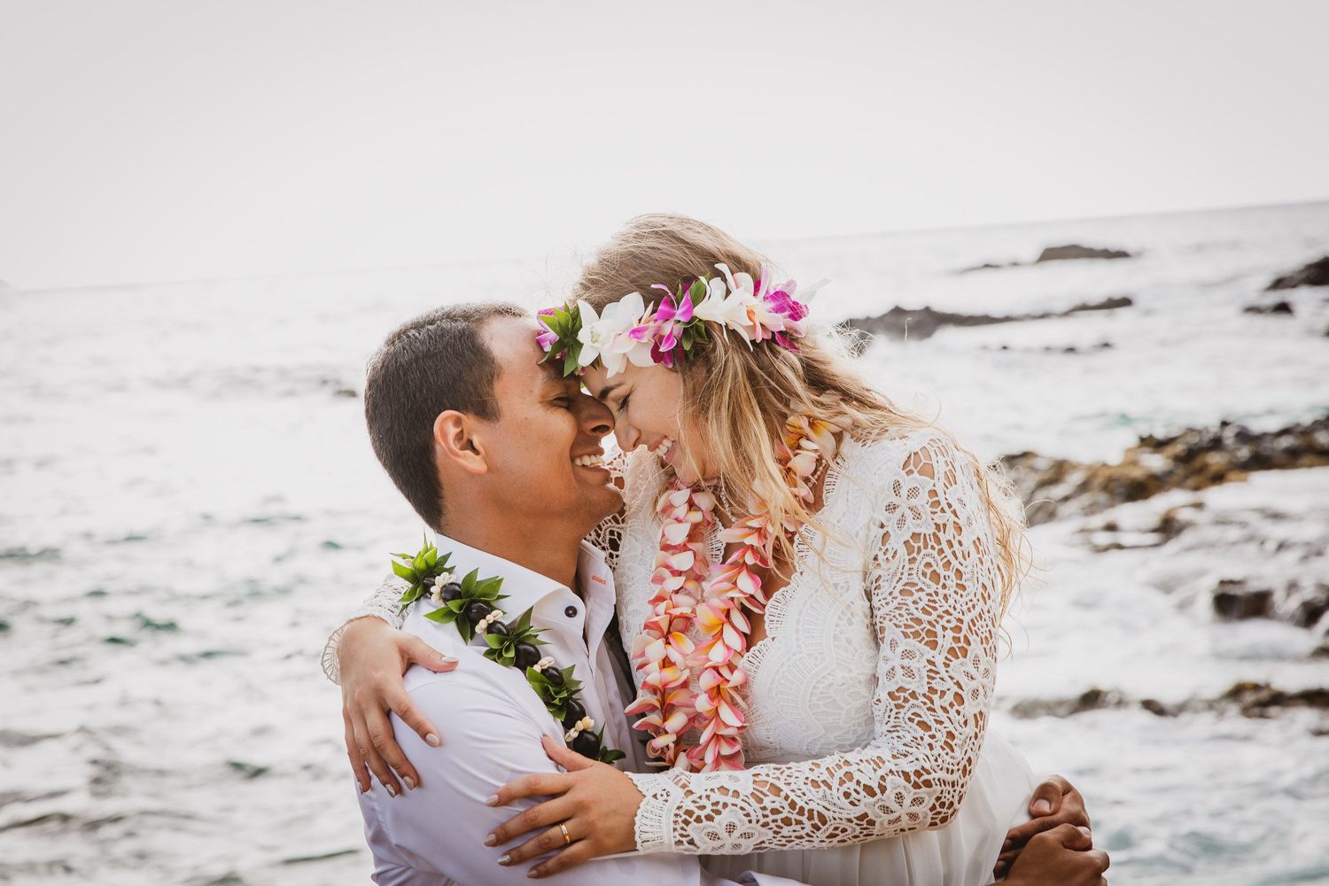 Couple sharing a romantic moment on rocky Hawaiian beach, bride in floral crown and lei, groom in white shirt.