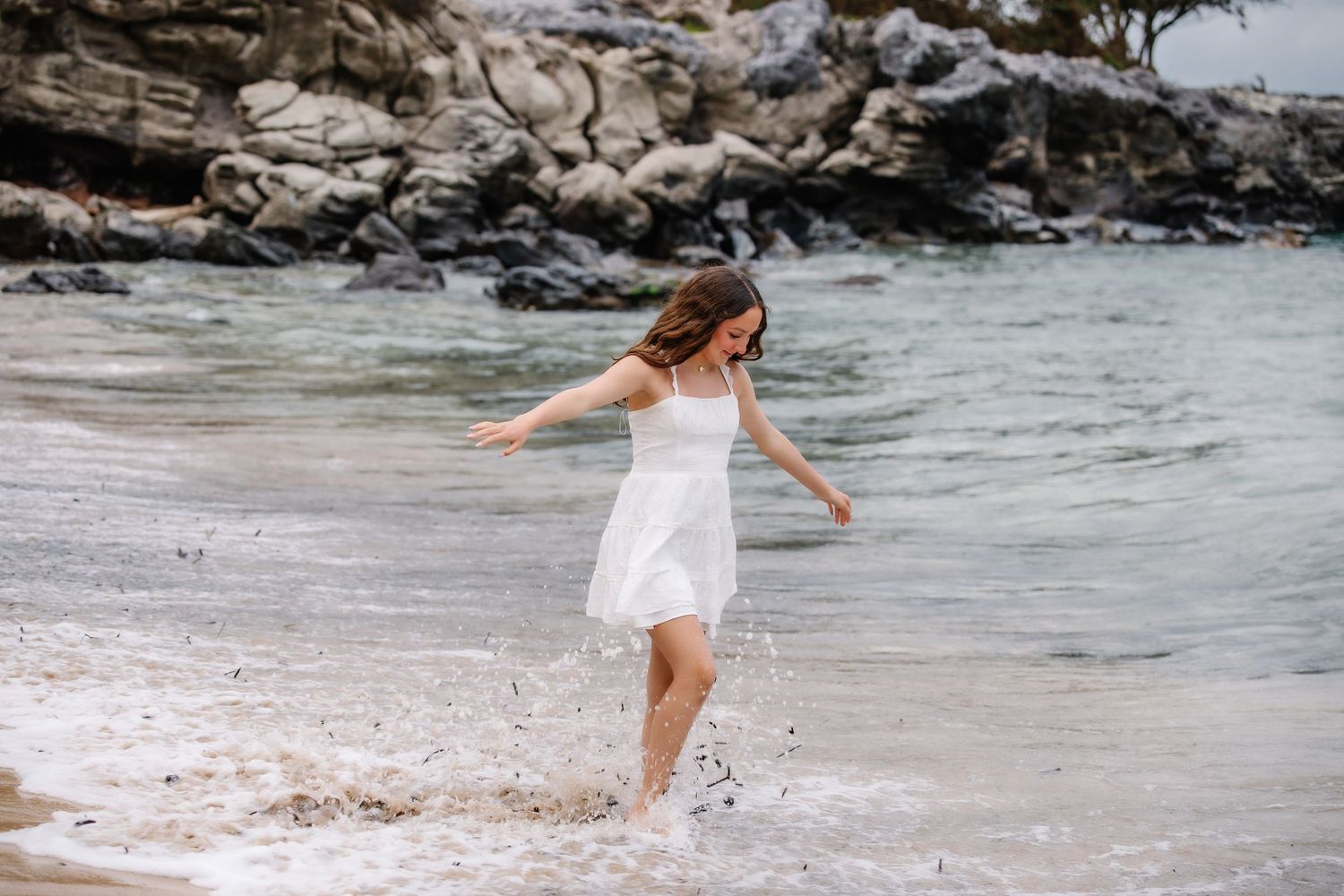 Young woman in white dress joyfully splashing in ocean waves on a rocky beach.