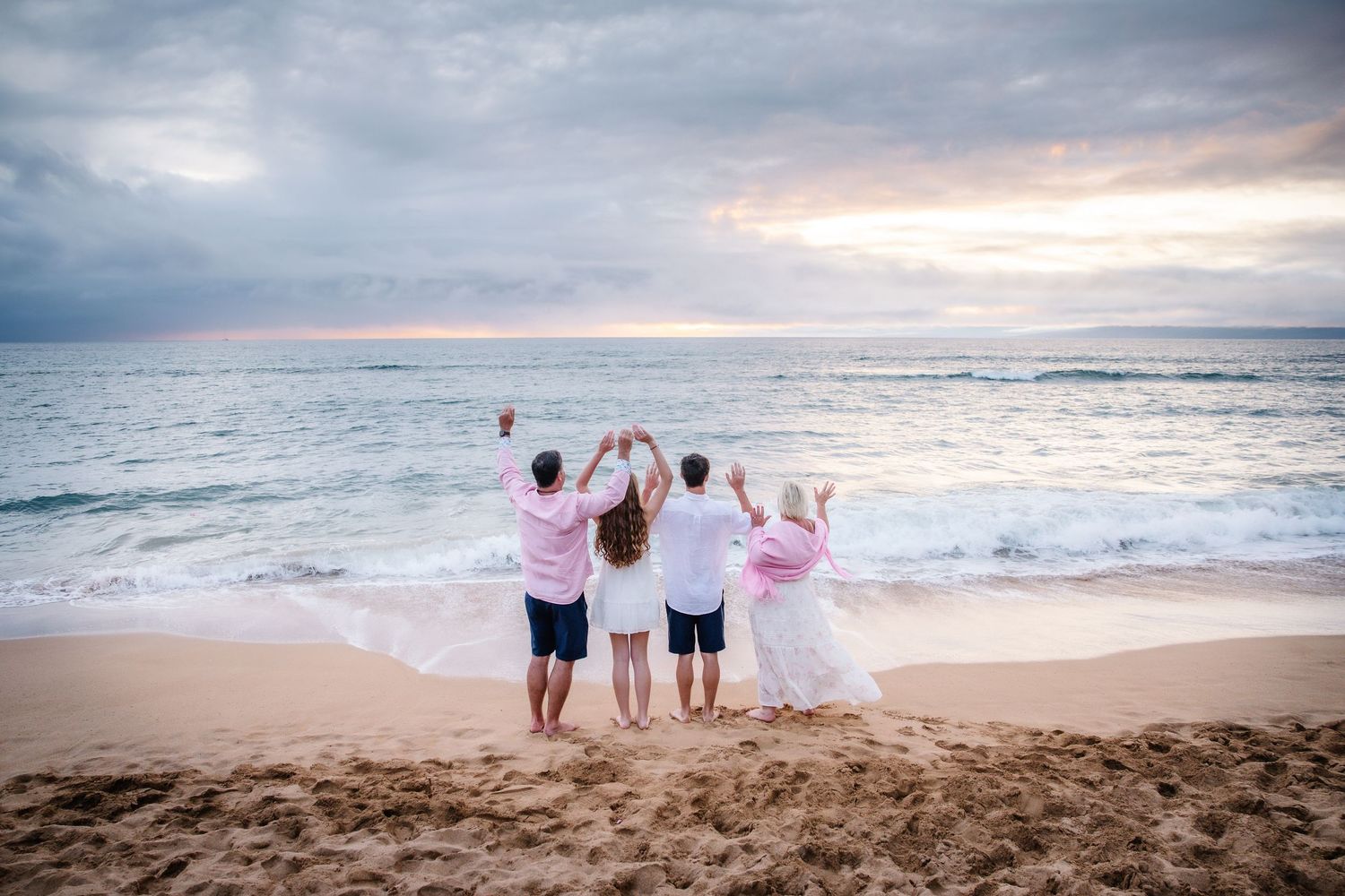Family of four celebrating on sandy beach with arms raised toward colorful ocean sunset.