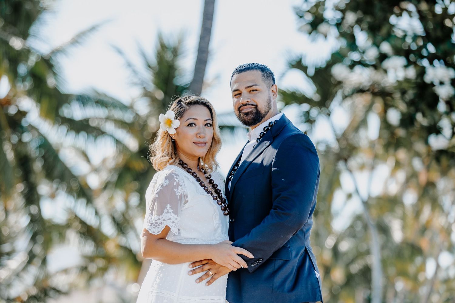 Couple in wedding attire posing outdoors with tropical palm trees, bride in white lace dress, groom in navy suit.